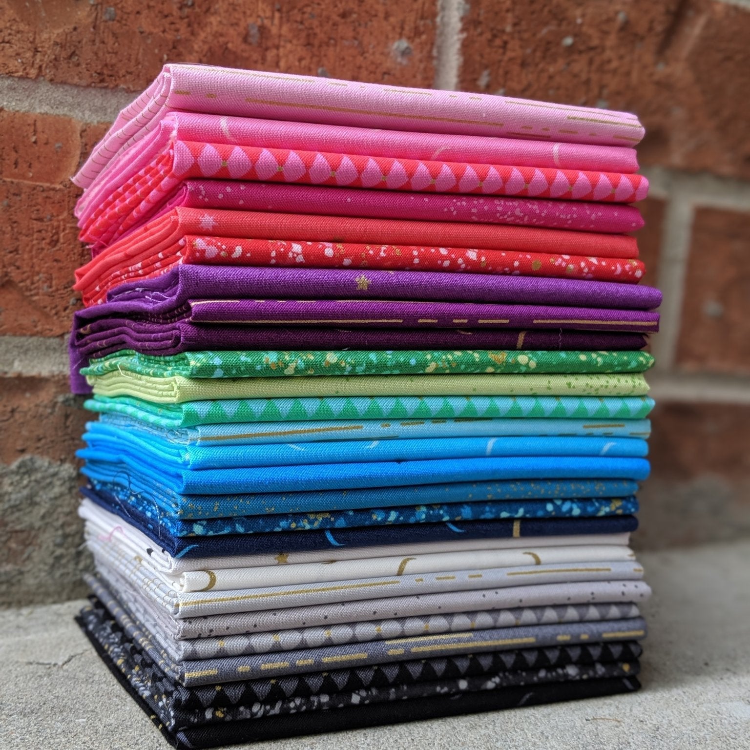 The image shows twenty-seven cuts of fabric folded on top of each other. Each fabric piece has its own color. Colors in this collection include black, gray, blue, purple, green, red, pink, and the other twenty colors are variants of those seven colors.