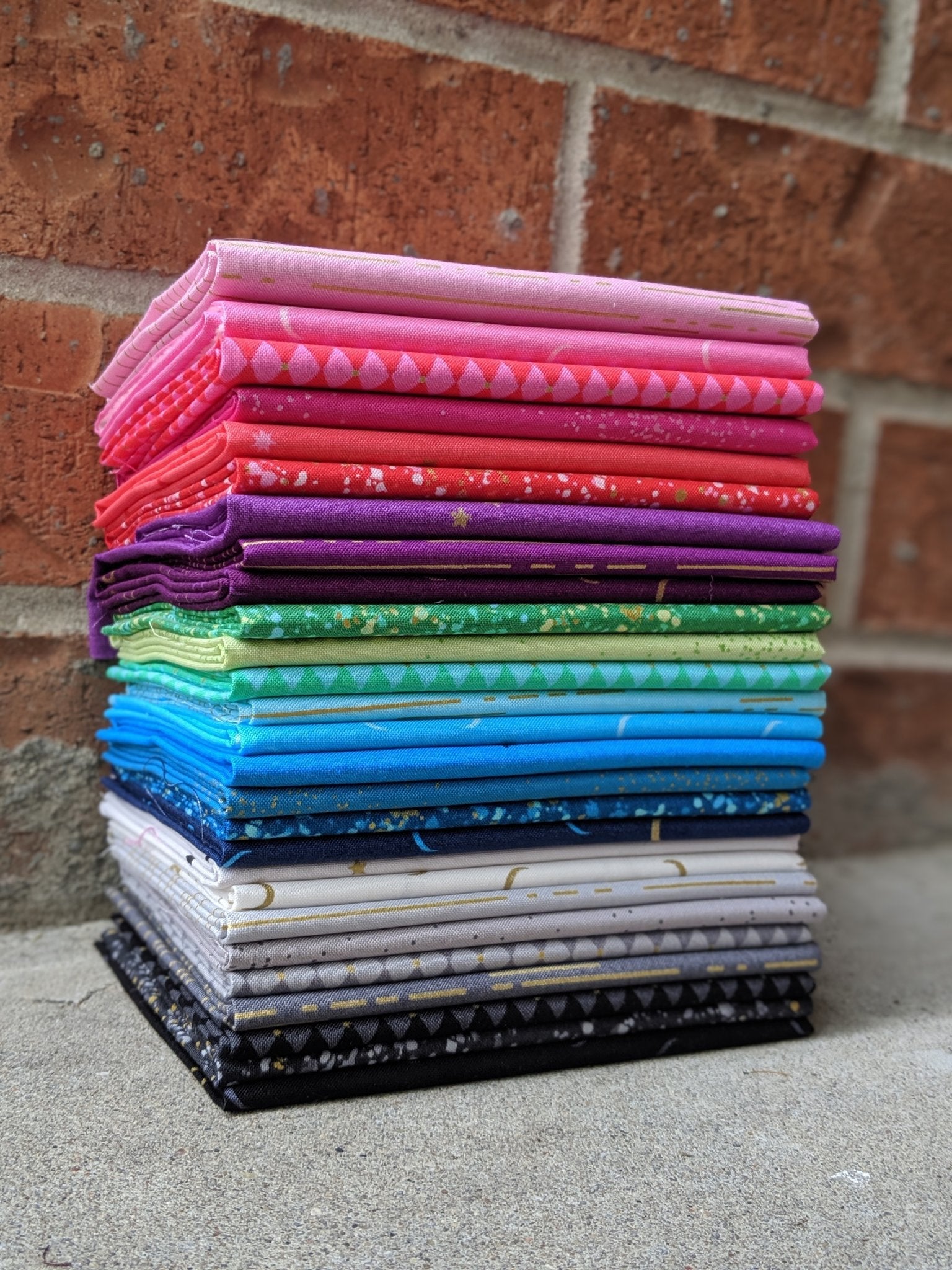 The image shows twenty-seven cuts of fabric folded on top of each other. Each fabric piece has its own color. Colors in this collection include black, gray, blue, purple, green, red, pink, and the other twenty colors are variants of those seven colors.