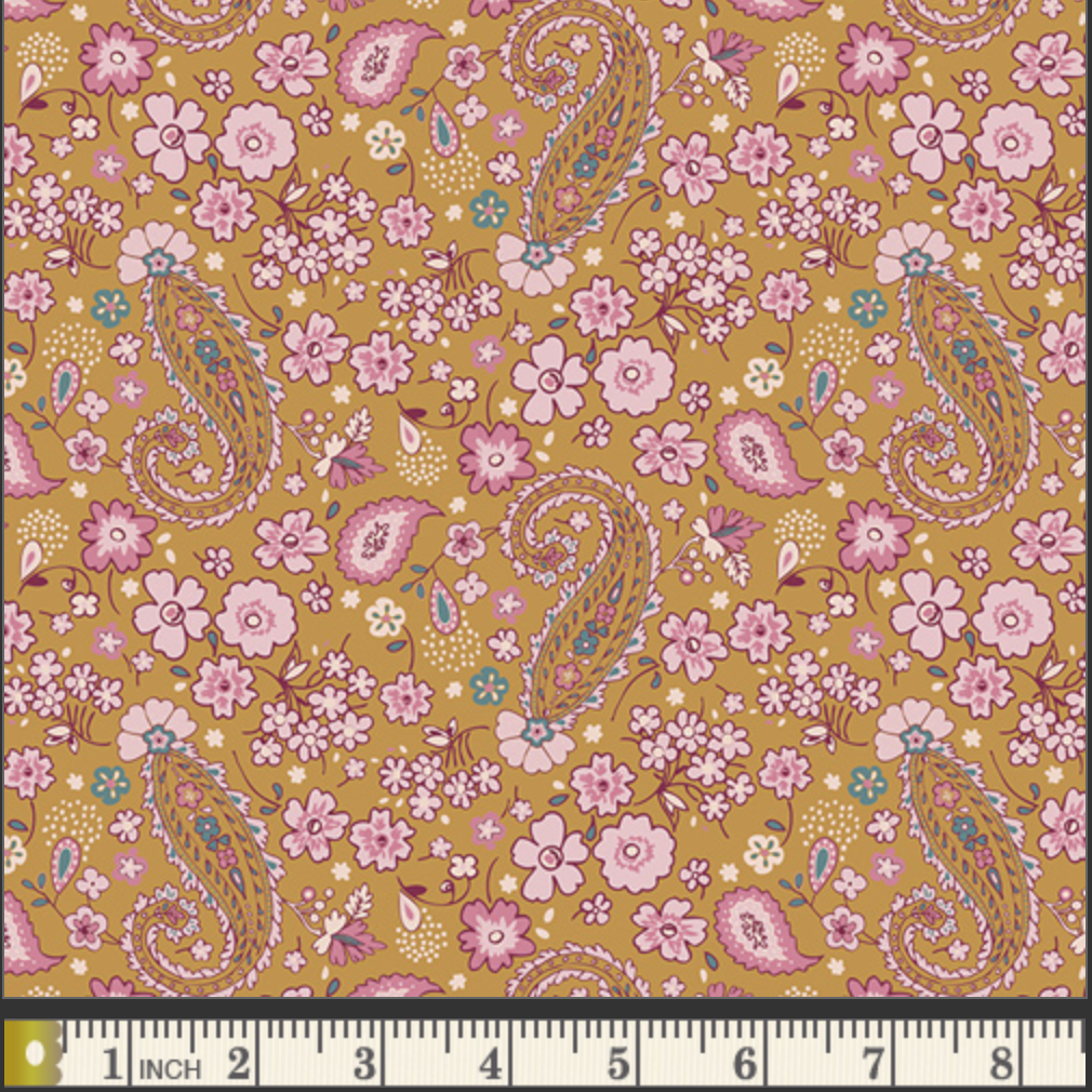A close-up of a floral and paisley patterned fabric sample on a mustard yellow background, with pink flowers, pink and blue paisley motifs, and a white inch ruler along the bottom edge for scale.