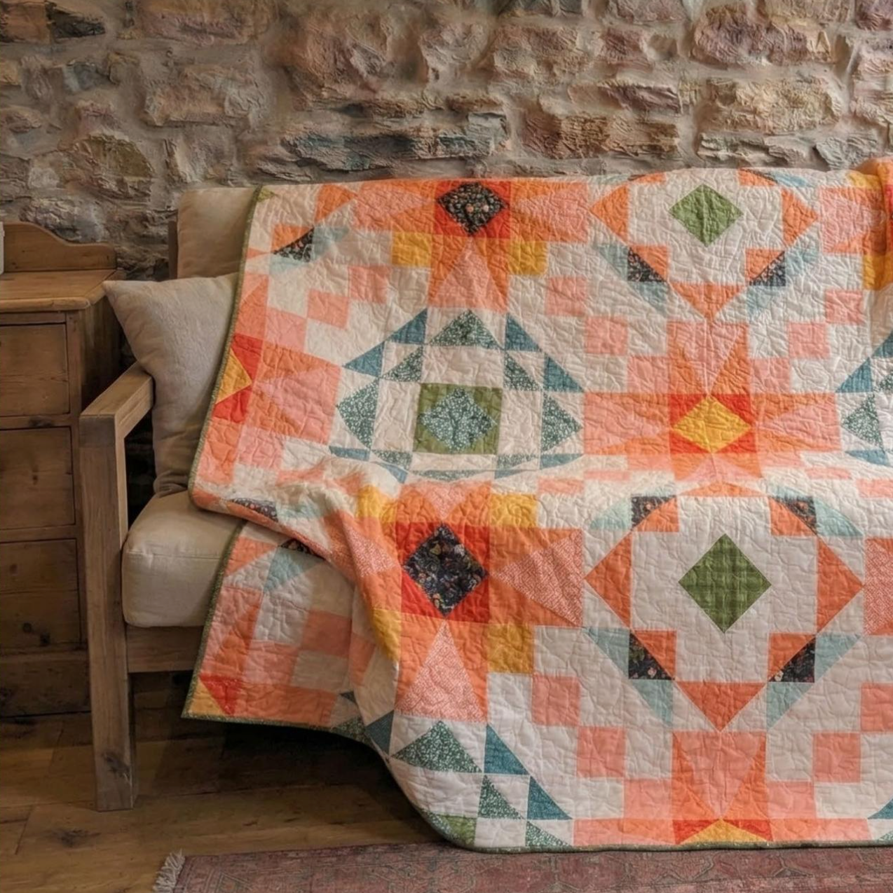 A multicolor geometric quilt draped over a rustic wooden chair in a cozy room with a stone wall background.